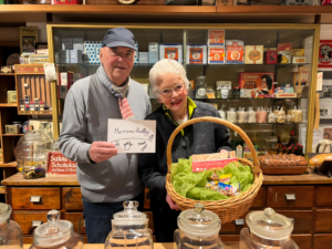 2 Personen vor einem alten Kaufmannsladen. In der Hand wird ein Korb gehalten.
