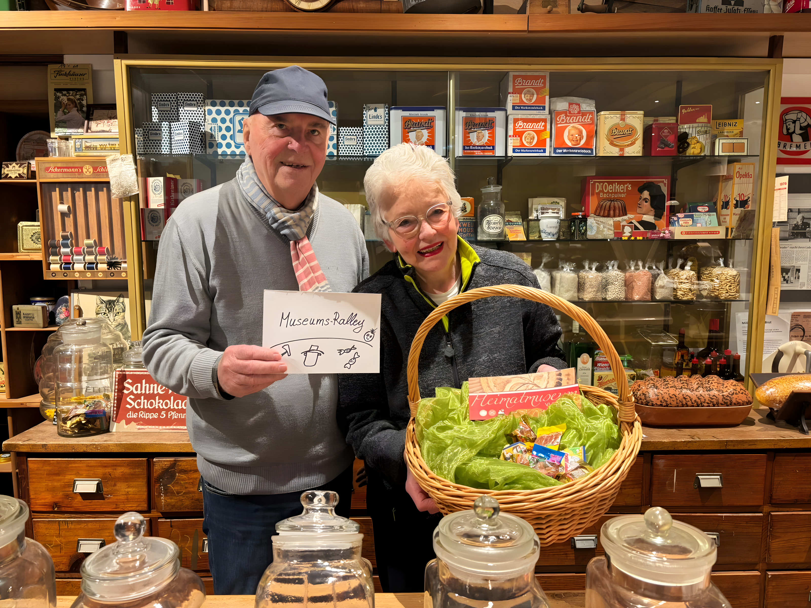 2 Personen vor einem alten Kaufmannsladen. In der Hand wird ein Korb gehalten.