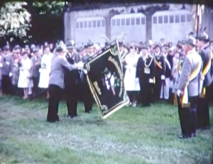 Viele Menschen in Schützenuniform mit einer großen Fahne in der Hand. Die Damen auch in festlicher Kleidung.